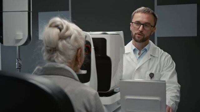 Medium shot of smiling male ophthalmologist using modern diagnostic machine while conducting eye exam for senior woman in clinic, copy space