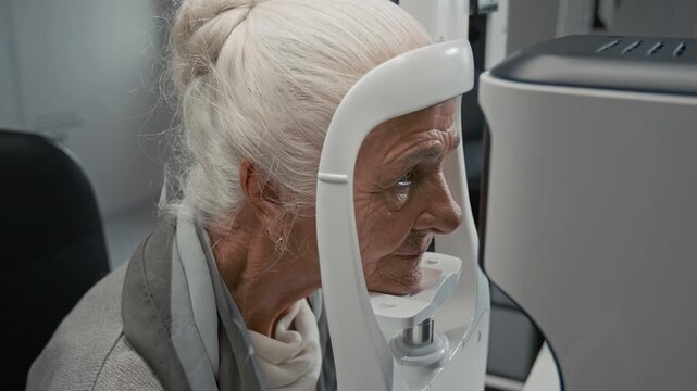 Close up shot of white haired senior woman putting head into frame of modern diagnostic machine during eye exam in ophthalmologist office, copy space