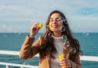 Woman blowing bubbles by the sea on a sunny day wearing a brown coat