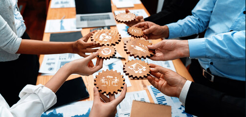 A diverse group of team members engages in a collaborative business meeting, holding wooden gears that represent management concepts and teamwork initiatives. Amity