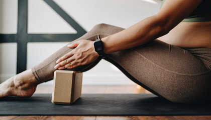 A person sitting on a yoga mat with a yoga block under their leg, wearing activewear and a smartwatch, in an indoor setting.