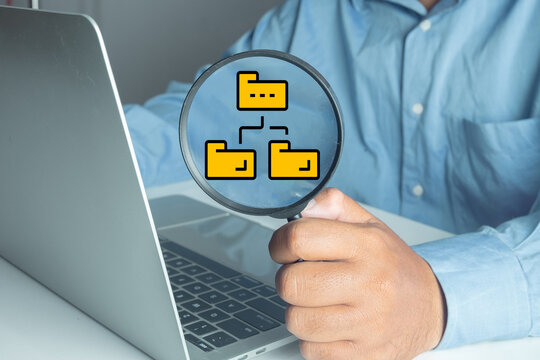 Hand holding magnifying glass focusing on yellow folder icons on laptop screen, representing data management, file organization, directory search, and digital storage concept in office. - Powered by Adobe