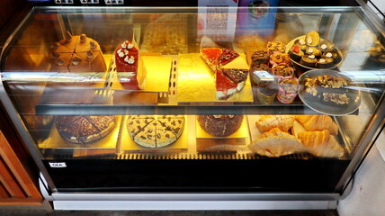Various cakes and desserts displayed in a glass refrigerated display case at a bakery, tempting...