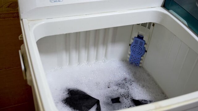 High-angle interior view of a semi-automatic washing machine violently agitating clothes during the wash cycle. Turbulent swirling water, foam, and motion blur inside the white tub.