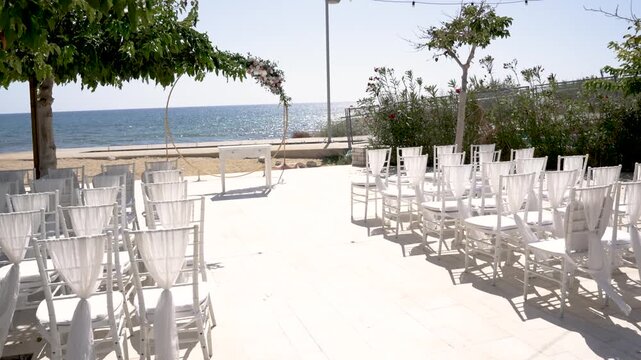 A beautiful setup for a wedding ceremony takes place on a sunny day by the beach. White chairs align facing the ocean, framed by greenery and a clear blue sky.