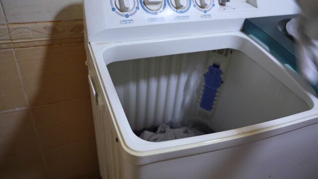 Close-up of a man's hand holding and dropping a purple shirt into an empty top-loading washing machine. Preparing for a domestic laundry wash cycle at home.