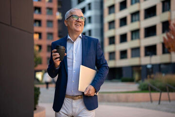 Happy senior businessman walking with coffee and laptop in city