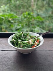 A bowl of stir-fried ferns on a table with a view of the green forest.