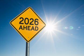 A yellow diamond road sign with the words 2026 ahead against a bright blue sky and shining sun