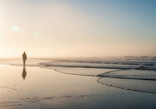 Lone figure walking on a wet beach shore during misty golden hour sunset with soft atmospheric reflection. - Powered by Adobe
