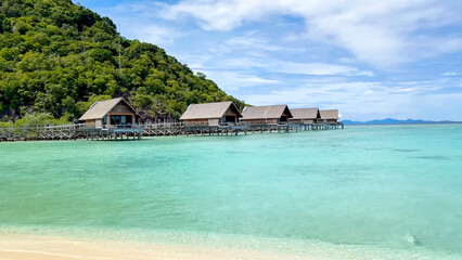 A secluded wooden beach bungalow sits near the water's edge, backed by a vibrant green hillside and turquoise ocean