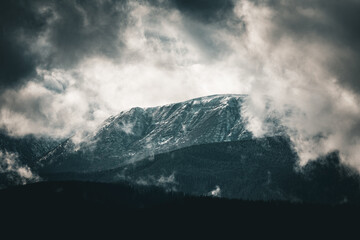 Moody, cool-toned clouds encircle a snowcapped mountain ridge above dark evergreen layers. Mist hangs over the forested slopes, creating a cinematic, alpine panorama with bold contrast and depth.