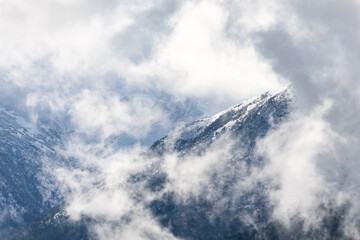 Snowy mountain peak breaks through swirling clouds and fog revealing rugged textures and winter detail. Ethereal alpine atmosphere evokes solitude, adventure and the dramatic power of wild nature.
