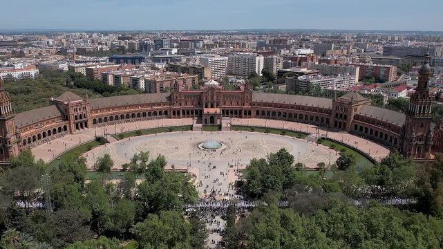 Wide zoom in aerial over Plaza de Espana in the Parque de Mar&iacute;a Luisa, Seville showing semicircular neoclassical Mudejar building, central fountain and visitors crossing sunlit expansive park square