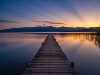 Fototapeta premium Stunning sunset pier view over serene lake waters with mountains, perfect for travel inspiration or peaceful mindfulness campaigns today