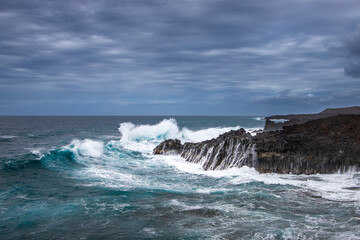 Rugged cliffs of Lanzarote's coastline are battered by powerful waves under a dramatic, stormy sky. The ocean appears turbulent, with shades of deep blue and white surf.