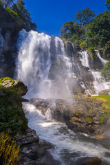 Stunning view of Wachirathan Waterfall in Chiang Mai, Thailand, with water crashing down over dark rocks and vibrant spray against a clear blue sky and green forest.