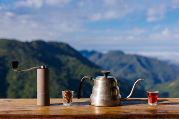 Coffee brewing set (kettle, hand grinder, beans) on a wooden deck, overlooking a magnificent mountain range under a beautiful blue sky on a clear morning.