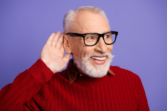 Happy senior man with grey beard and glasses smiles as he listens closely against a purple background