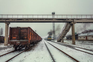Winter train station with rusted freight cars and steam locomotive under a concrete bridge in snowy landscape