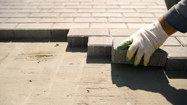 Construction worker hands wearing gloves laying gray concrete paving stones on sand base for a new sidewalk or patio