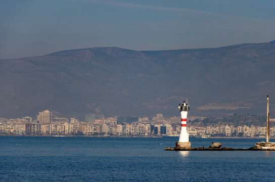 Iconic Red and White Striped Lighthouse and Old Beacon Tower Standing in the Calm Sea, Set Against a Hazy Mountain and Densely Built Coastal City Skyline.