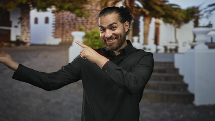 Man points finger to left at white building steps outdoors, showing bare hand and smiling in black shirt, casual young adult; cheerful.