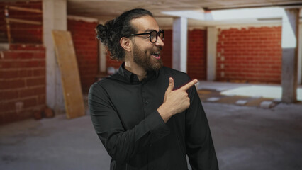 Obraz premium Man in black shirt pointing right and giving thumbs up inside a building construction site; confidence success.
