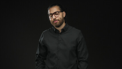 Obraz premium Man with glasses and beard, eyes closed and head tilted, hands in pockets wearing a dark shirt in a black studio; contemplative.