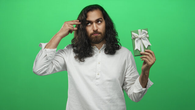 Man with long hair and beard in white shirt holding a green wrapped gift with white bow and making finger to temple gesture in green studio; wry confusion. - Powered by Adobe