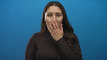 Young hispanic woman with long straight dark hair covers her mouth with right hand in blue studio; surprise.