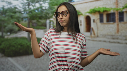 Woman holding palms up in a shrug gesture with glasses and striped shirt on a street by a stone...