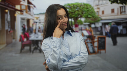 Woman points finger to cheek with arms crossed on a restaurant terrace near sidewalk tables, facing camera and smiling slightly; playful teasing confidence.