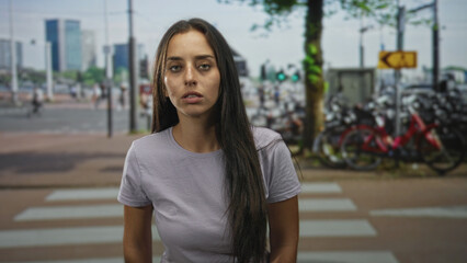 Woman standing at street crosswalk with visible face acne and long hair, looking down amid parked bicycles and city buildings; pensive solitude.
