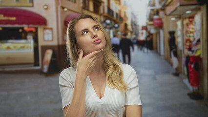 Fototapeta premium Woman thinking on lively city street with blonde hair, wearing white shirt, surrounded by shops and urban environment, reflecting an outdoor lifestyle.