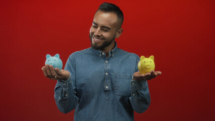 Young hispanic man holding blue and yellow piggybanks with both hands in red studio; saving choice...