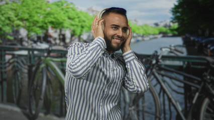 Man holding headphones to ears on street by amsterdam canal, smiling and wearing striped shirt next to parked bicycles on bridge; joy music escape.