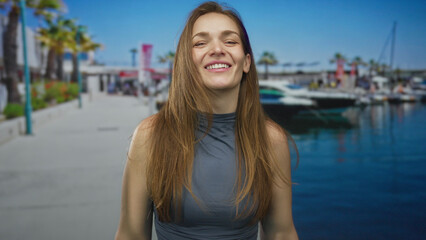 Young woman finger pointing at camera on sunlit port dock near moored boat under blue sky; confidence.