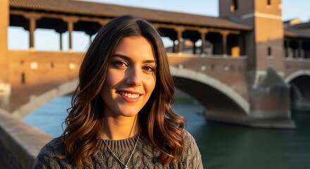 Smiling woman with long brown hair stands on a bridge at sunset.