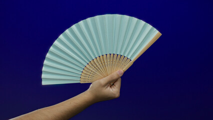 Hand holding blue fan against isolated blue wall background with a man's arm visible, emphasizing...