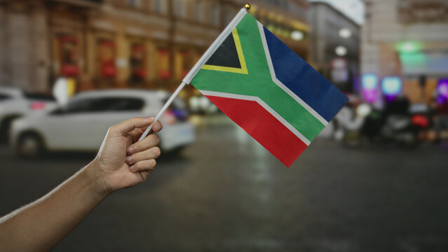Hand holding south african flag in city street capturing dynamic cultural essence in urban setting during evening with diverse people and lights in background.