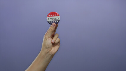 Hand holding a patriotic button reading 'i voted' against a solid purple background symbolizing democratic participation and civic duty.
