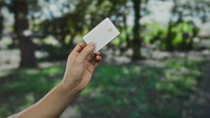 Fototapeta premium Man holding white credit card in outdoor park setting with lush green background, showcasing finance and technology concept in a natural environment.