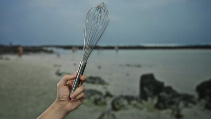 Man holding whisk at seaside beach, capturing outdoor culinary creativity with a caucasian male hand against a serene coastline backdrop.
