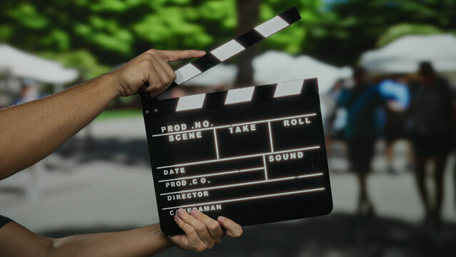 Man holding clapperboard in outdoor market with blurred people in background on a sunny day.