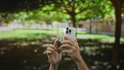 Man holding smartphone capturing photo in sunny park with lush green trees and bright light filtering through leaves showcasing outdoor technology interaction and nature backdrop.