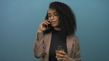 Woman with curly hair in glasses holding coffee cup and talking on smartphone against isolated blue wall background, conveying business communication.