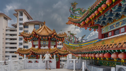 A man stands on the terrace of a Chinese temple, leaning on a stone railing. Curved roofs of pagodas, rows of red lanterns along the edges of the cornices. A dragon figure against the sky. Malaysia.