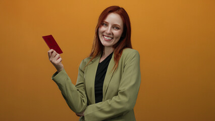 Woman with red hair joyfully holds a norwegian passport against an isolated orange background, highlighting personal identity and global travel. © Krakenimages.com