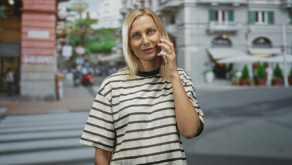 Woman holds phone to ear at street crosswalk near cafe awnings and city buildings, striped shirt visible; calm conversation.
